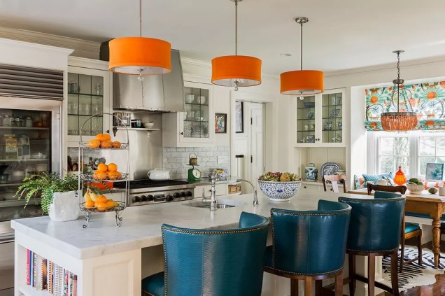 Para animar este espaço de cozinha neutro, o escritório Katie Rosenfeld Interior Design empregou vários tons de verde, azul e laranja. Tecidos estampados e bonitos alegram o recanto do café da manhã. Três tons de laranja colorem as luminárias e três bancos de bar azuis complementam a ilha da cozinha.