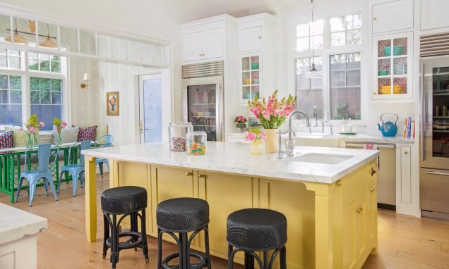 Uma ilha amarela ancora esta cozinha da designer de interiores Alison Kandler. Os armários superiores revestidos com papel de parede rosa choque separam o espaço em branco. Móveis verdes e azuis combinados com travesseiros vigorosos criam um recanto extra para o café da manhã.