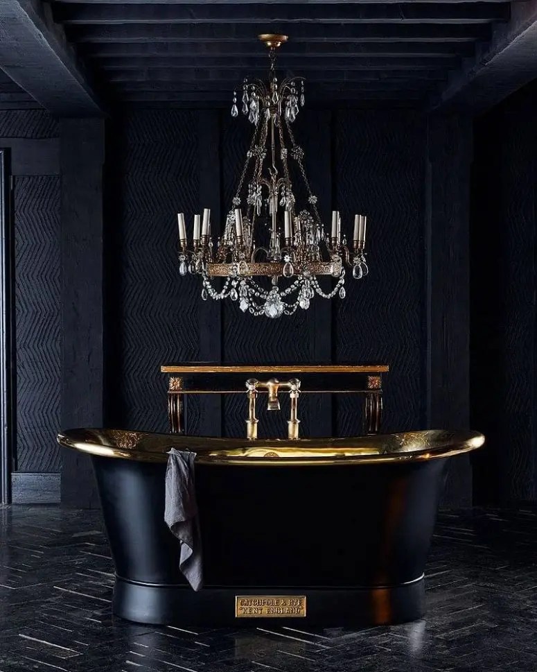 Banheiro refinado com paredes cinza grafite textural, uma banheira polida preta e dourada, um lustre de cristal e um piso de parquet preto.