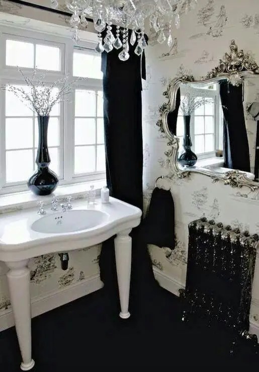 Um banheiro gótico preto e branco com papel de parede, uma pia com pernas, um lustre de cristal e um belo espelho em uma moldura ornamentada.