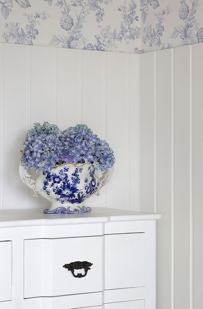 Floreira de porcelana branca com flores azuis pintadas. Hortênsias arranjadas dentro dela. Ao fundo, papel de parede com flores azuis.