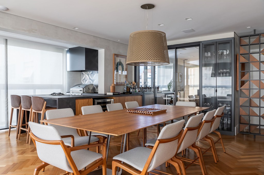 Sala de jantar, com mesa de madeira e dez cadeiras. Ao fundo, a área da churrasqueira