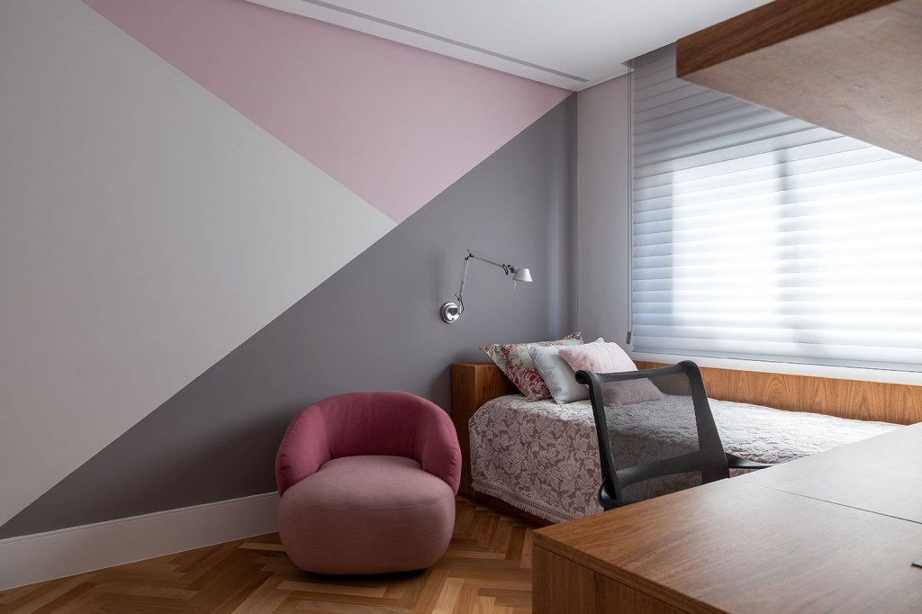 Quarto com três triangulos coloridos na parede, poltrona rosa, cama com roupa rosa e mesa de madeira