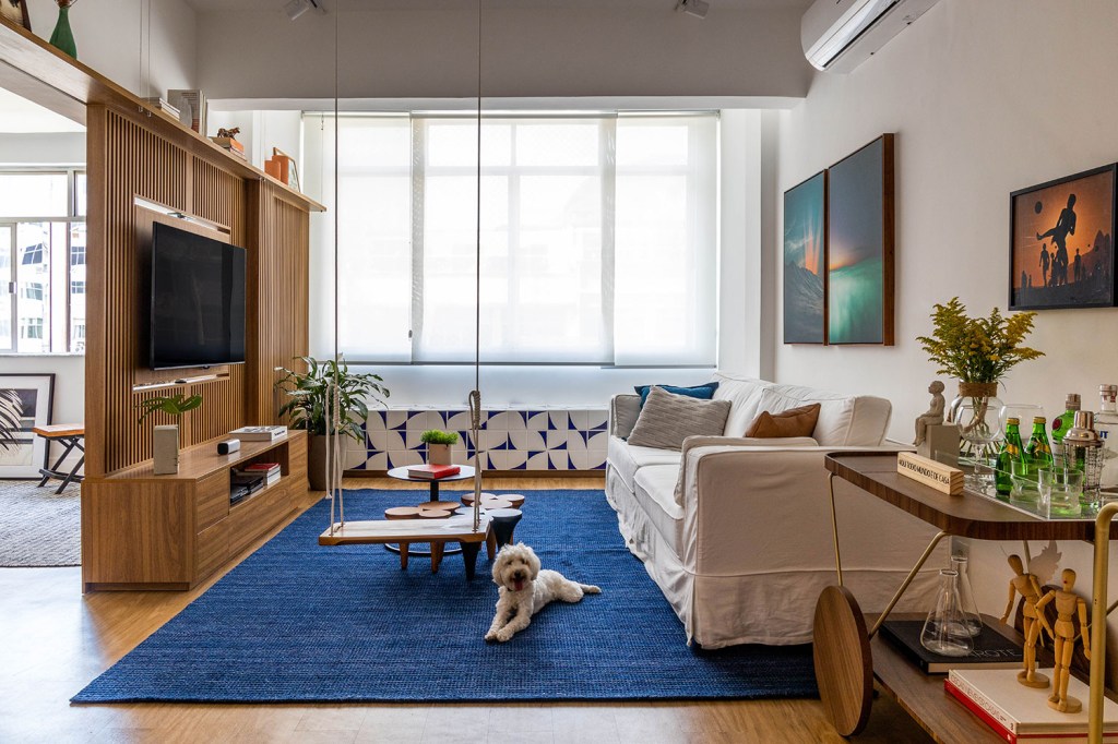 Sala de estar com tapete azul, TV sobre um painel ripado, sofá branco e tapete azul, com uma cachorrinha branca deitada sobre ele