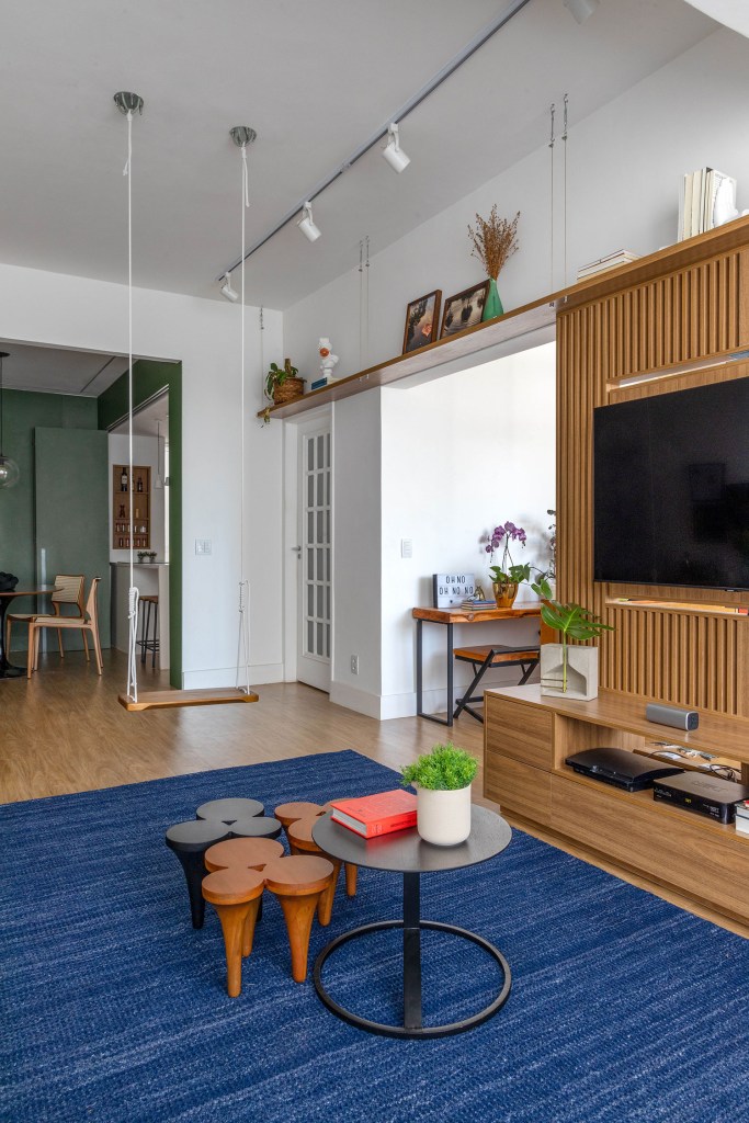 Sala de estar com movel de centro sobre tapete azul e ao fundo, a sala de jantar com mesa redonda com quatro lugares e paredes verdes e visão parcial para cozinha