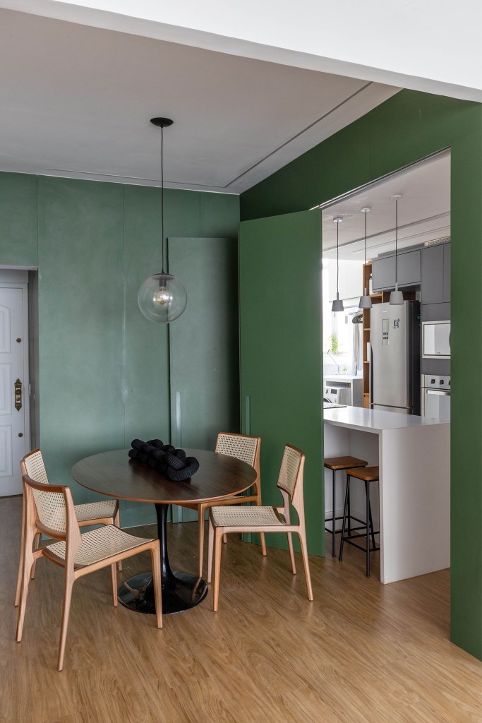 Sala de jantar com mesa redonda com quatro lugares e paredes verdes e visão parcial para cozinha