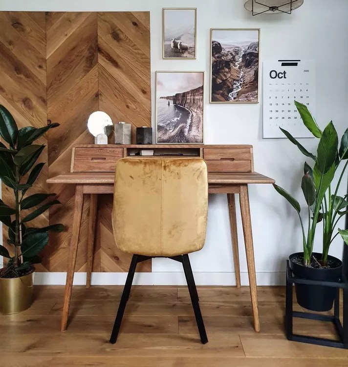 Home office com mesa de escritorio pequena, com piso de madeira e detalhe em madeira com paginação escama de peixe na parede junto de quadros