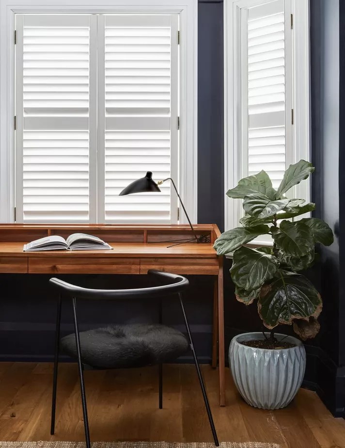 Home office com janelas brancas, mesa de madeira e planta