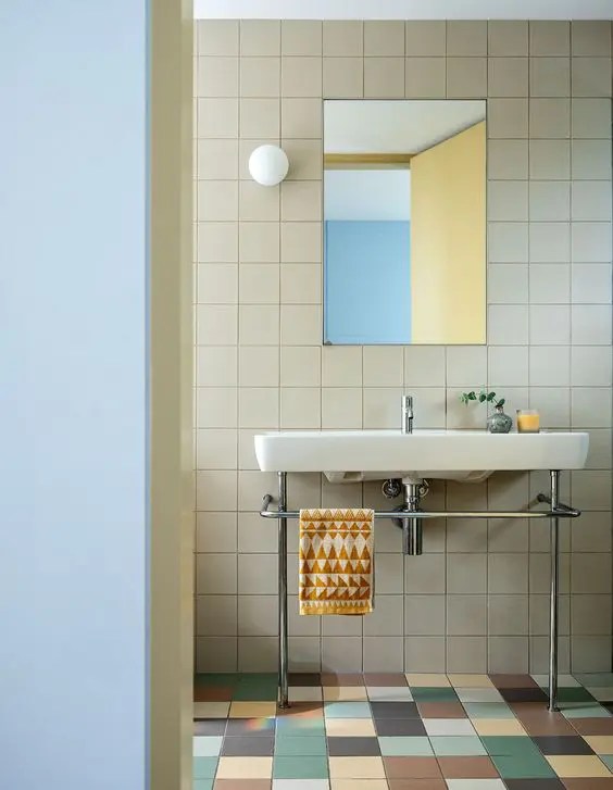 Um cômodo moderno revestido por peças neutras na parede e coloridas no chão, com um lavatório independente e um espelho retangular
