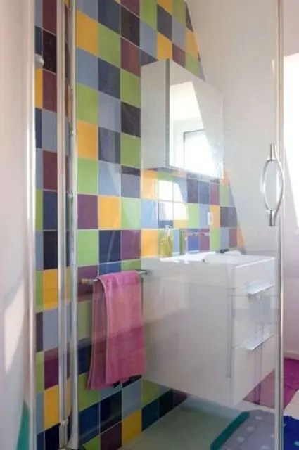 Uma parede de azulejos de cores aleatórias, um pequeno espaço de chuveiro e uma penteadeira branca flutuante