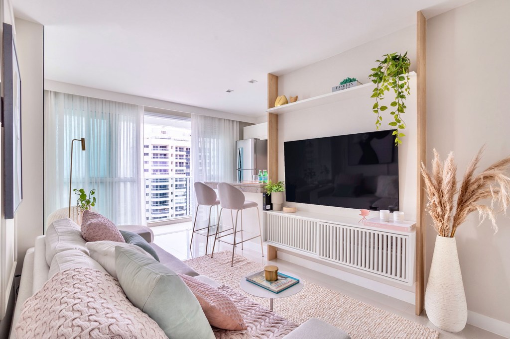 Sala de estar com varanda integrada, com sofa branco com almofadas cinza e rosa. Na frente, uma TV em painel com prateleira acima com objetos decorativos