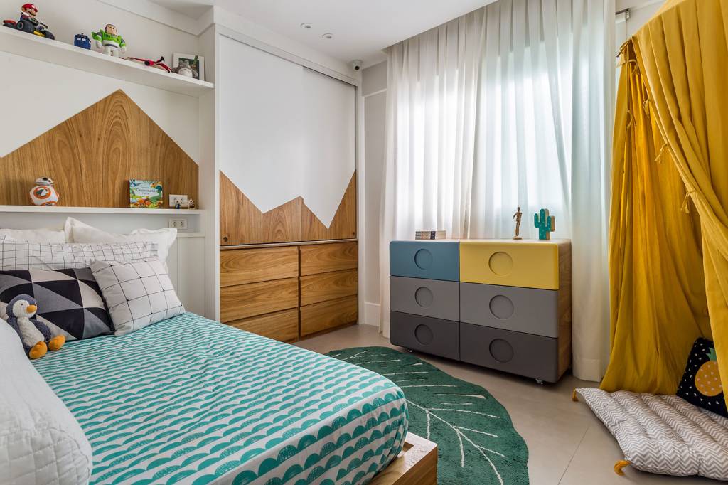 Quarto infantil, com roupa de cama verde, tapete em fomarto de planta, guarda roupa com desenho de montanha, e cômoda nas cores cinza escuro, cinza claro, azul e amarelo