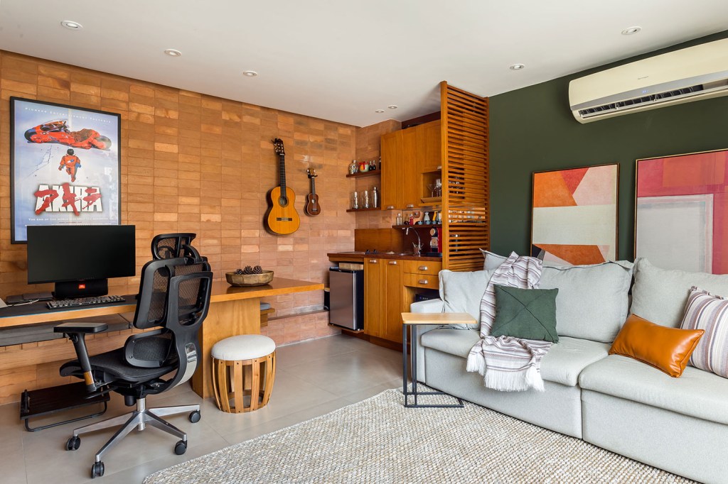 Sala e home ofccies com paredes de tijolo, violão pendurado, estantes de madeira e sofá cinza em frente a uma parede verde com quadro