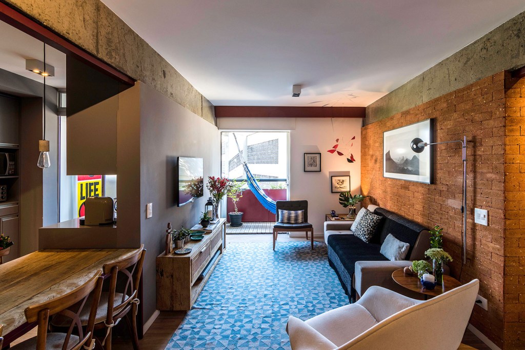 Sala de estar integrada, com piso de madeira, parede de tijolinhos, ao fundo, varanda com rede