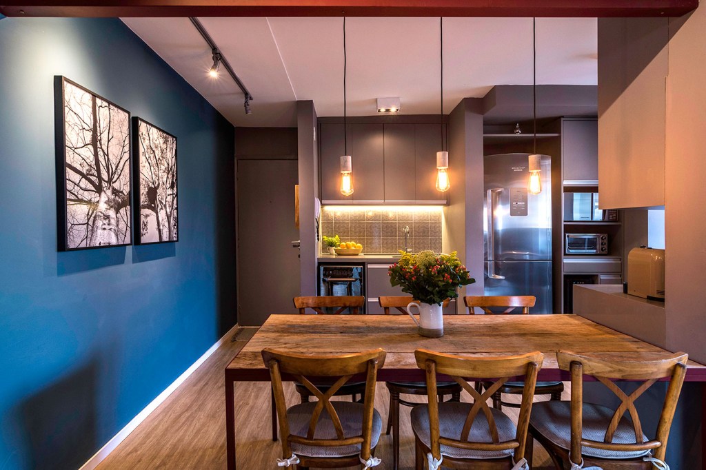 Sala de estar jantar, com piso de madeira, e cozinha com móveis marrons e aprede azul à esquerda