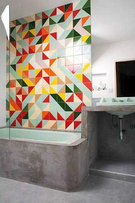 Uma parede moderna com azulejos geométricos super brilhantes combinando com o concreto e a banheira verde, que tomam conta do espaço