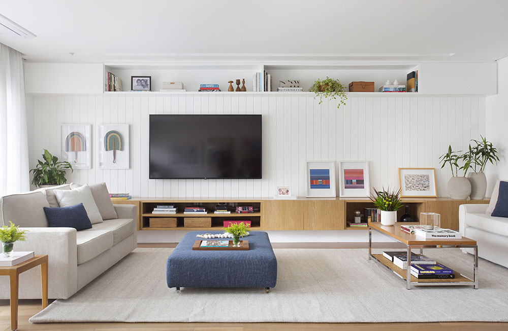 Living com sofá branco, rack em madeira, tv e pufe azul como mesa de centro