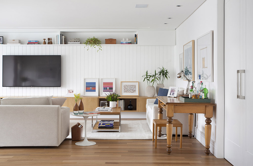 Sala com piso em madeira, sofá claro e painel em madeira branca no fundo com tv