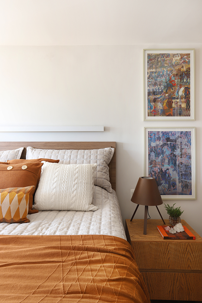 Quarto com cama de casal e almofadas alaranjadas. Quadro na parede. Mesinha lateral em madeira com abajur
