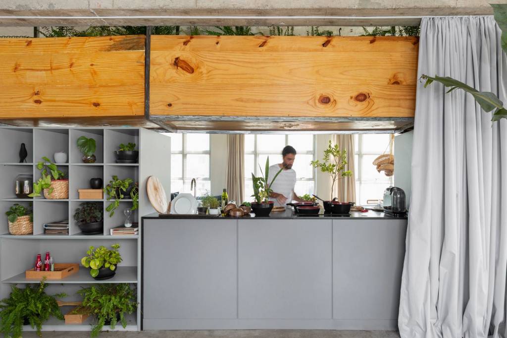 arquiteto-transforma-sala-comercial-em-loft-julia-ribeiro-cozinha-aberta-cortina-blackout-estante-com-plantas Cozinha compacta com estantes que acomodam plantas ao lado e caixas de madeira que são vasos de plantas.