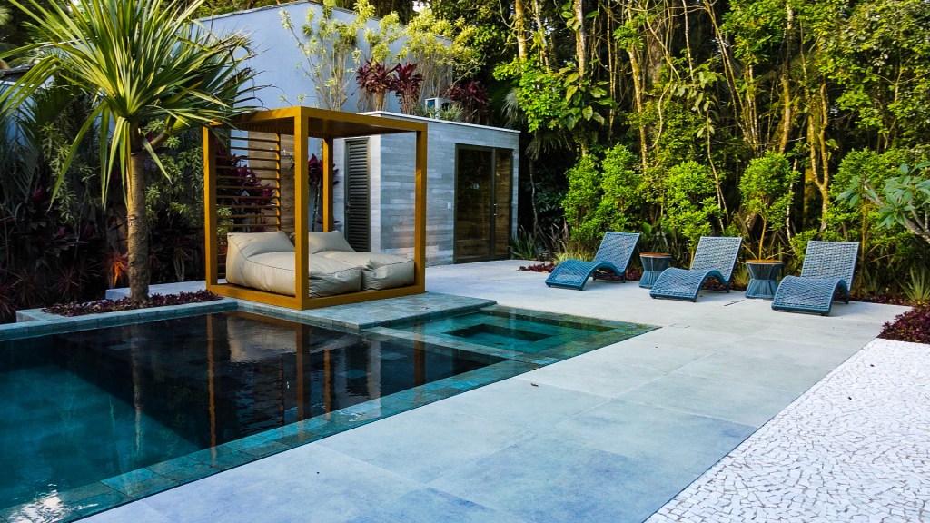 Detalhe da piscina com espelho d'água e espreguiçadeiras e móveis externos sob pergolado, com jardim tropical ao fundo.
