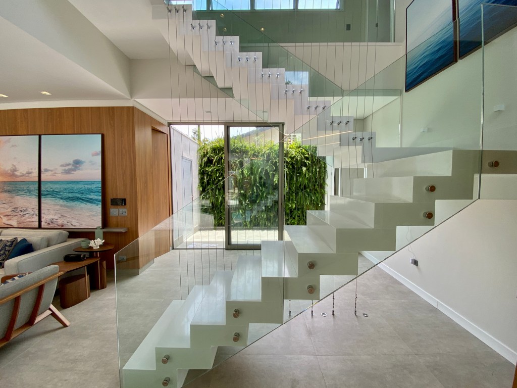 Escada branca com guarda-corpo em vidro em living amplo com paredes revestidas de painéis de madeira.