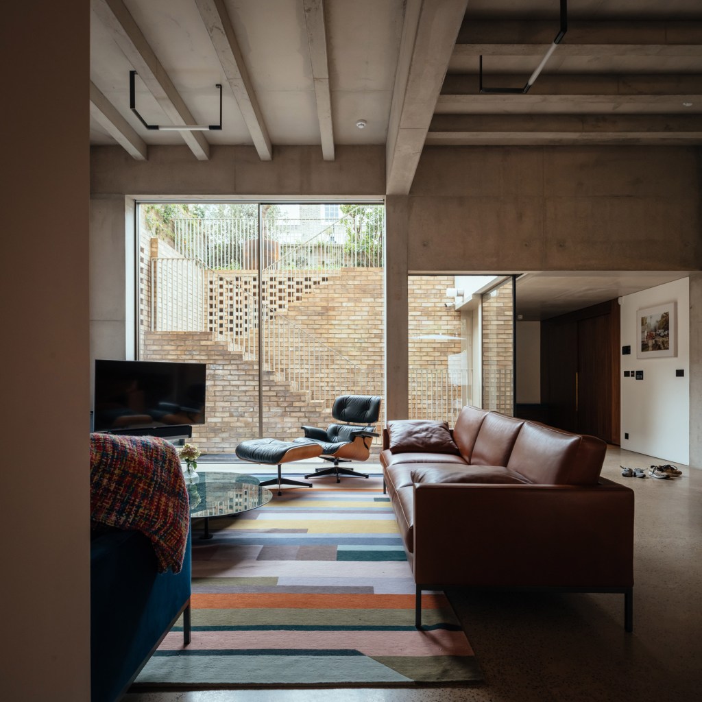 Sala de estar com sofá de couro grande, tapete colorido geométrico, teto em concreto aparente e aberturas de vidro que oferecem luz natural