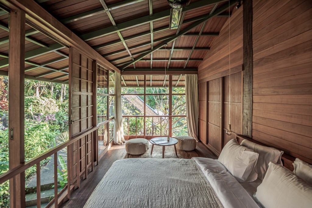 Quarto rústico em madeira com cama de casal e janelas de vidro que integram à paisagem do jardim.