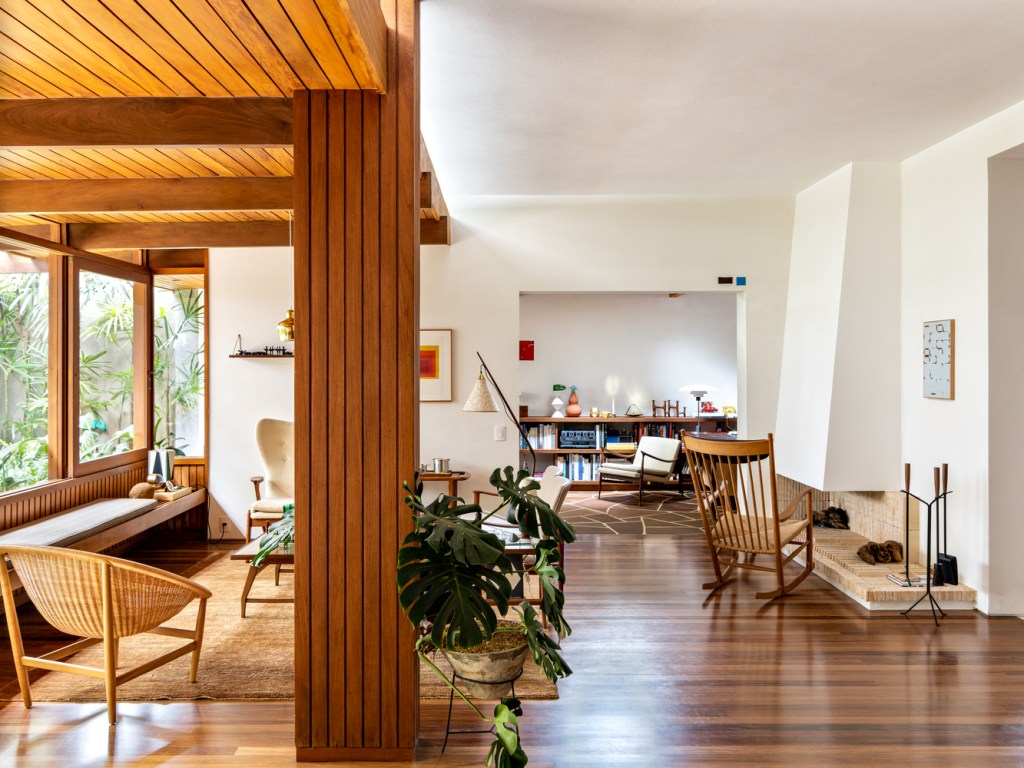 Sala de estar com piso de madeira corrida, mobiliário modernista, lareira embutida na parede e coluna revestida de madeira em primeiro plano.