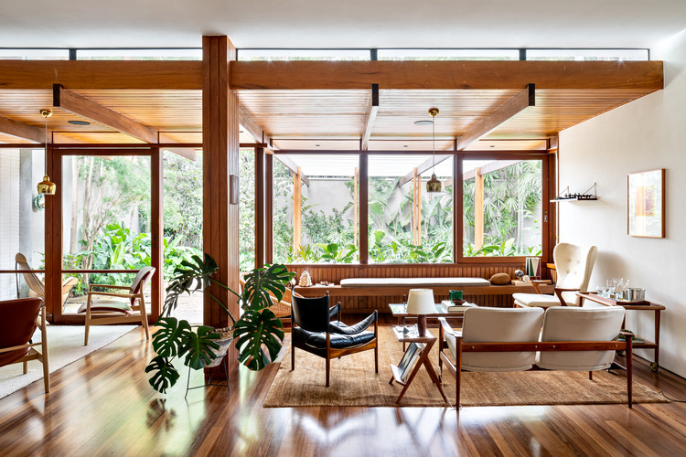 Imagem mostra sala de estar com piso de madeira, mobiliário modernista, aberturas de vidro com caixilhos de madeira e integração visual com o jardim externo.