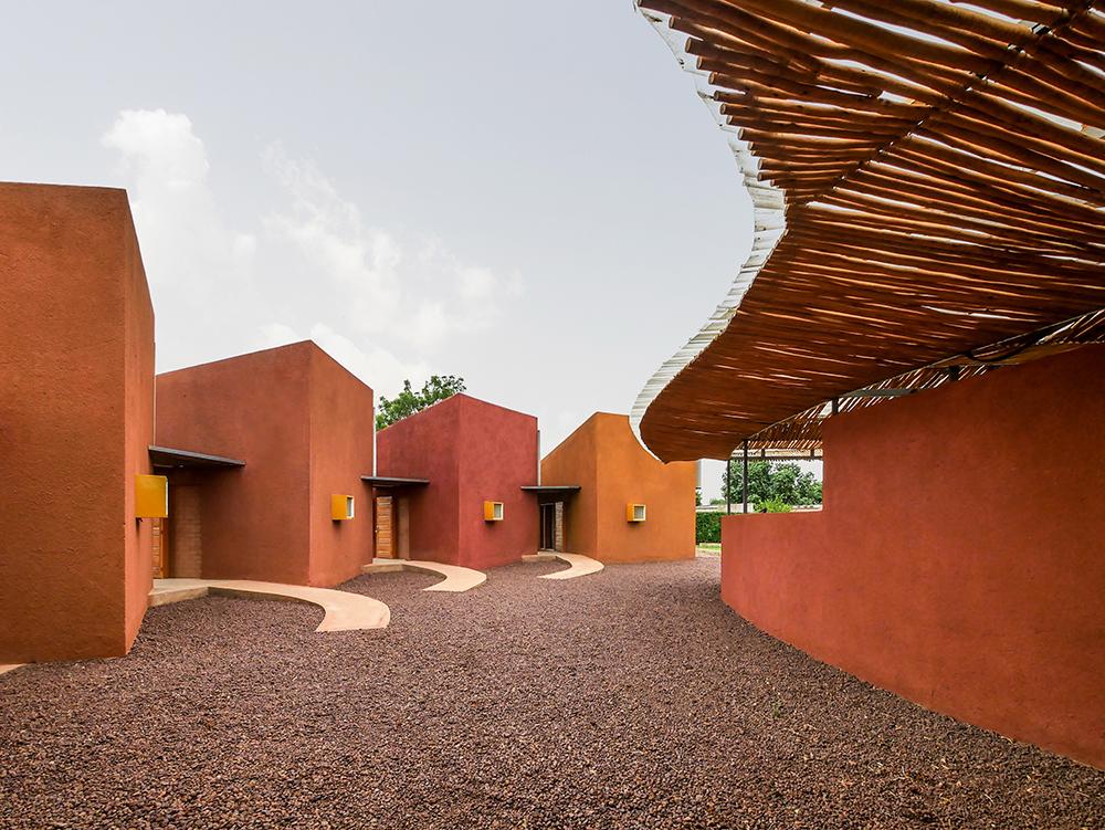 Imagem mostra fachada do projeto Leo Doctors Housing, construção de terra pintada em cores naturais, chão de pedras e cobertura de bambu.