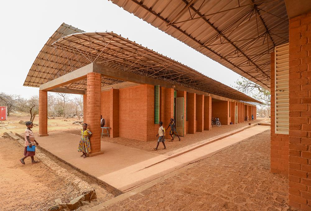 Imagem mostra prédio horizontal com tijolos aparentes, cobertura com telhas metálicas e algumas crianças em frente, em paisagem árida africana