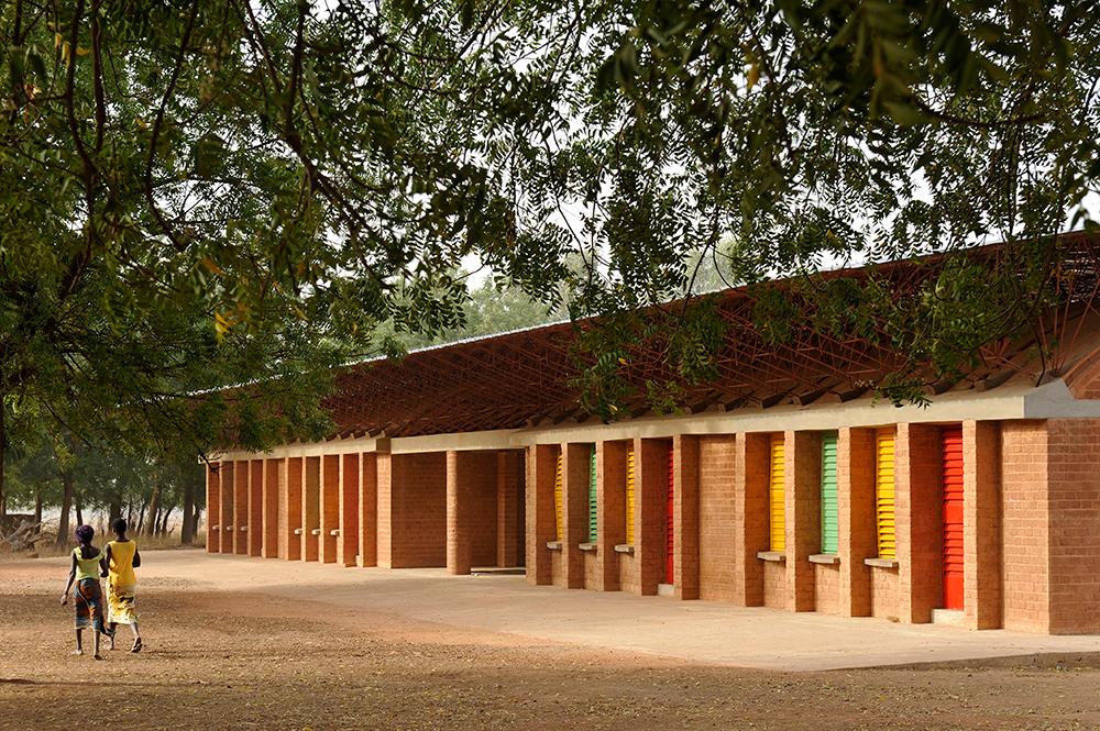 Imagem mostra árvore grande e fachada da escola em Gando, construída com tijolos aparentes e janelas coloridas em amarelo, verde e vermelho.