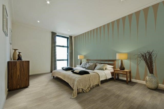 quarto de casal com cama, duas mesas de cabeceira com abajures, parede verde com desenhos geométricos e piso vinílico em padrão amadeirado.