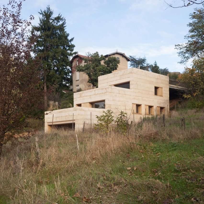 Casa semienterrada em paisagem bucólica de floresta tem fachada em tom de areia.