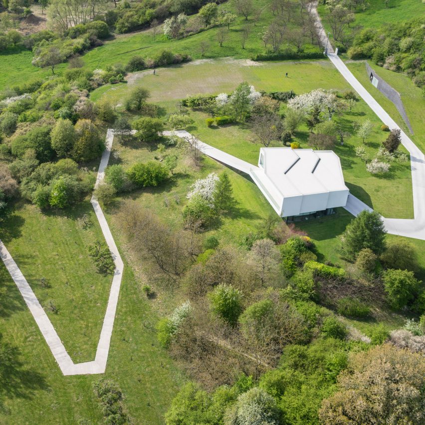 Foto aérea mostra estrada de concreto branco em meio à paisagem verde, com uma casa inteira na cor branca.