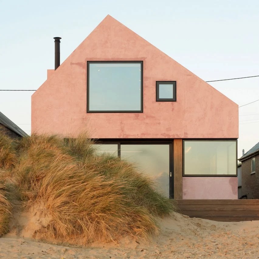 Casa com telhado triangular e fachada rosa, com chaminé e aberturas em vidro, na praia.