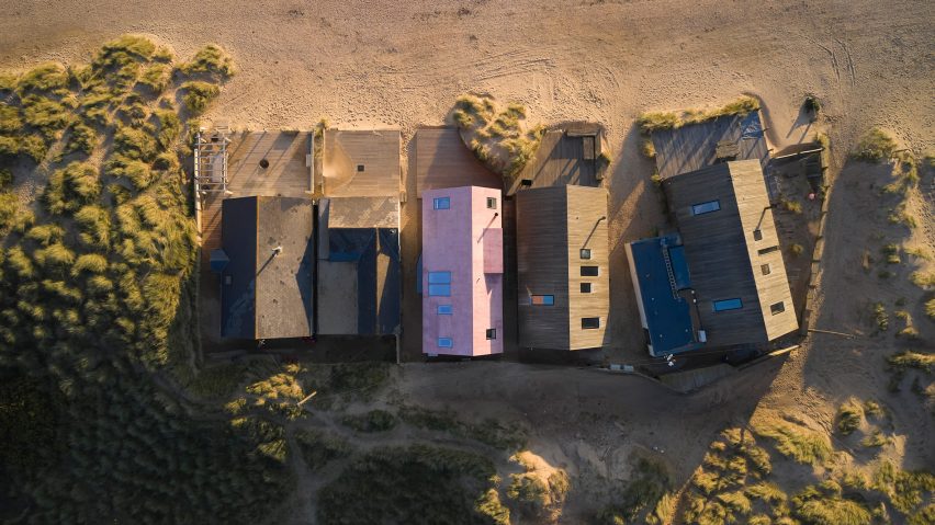 Foto aérea mostra cinco casas em frente à praia, sendo a do meio com telhado rosa que se destaca entre as demais.