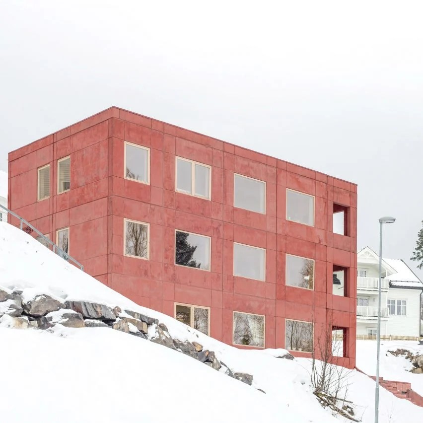 Prédio com dois andares e fachada em concreto em tom vermelho e janelas quadradas de vidro, em paisagem com neve.
