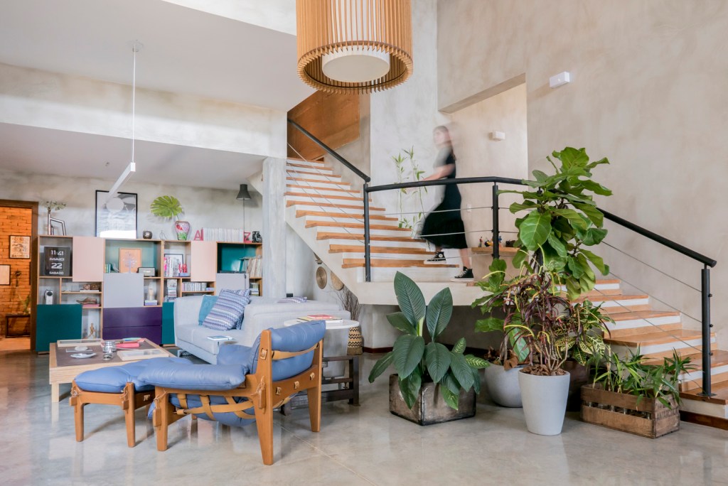 Imagem mostra sala de estar com móveis coloridos, como a poltrona mole de Sérgio Rodrigues, além de uma escada para o piso superior e diversos vasos com plantas tropicais.