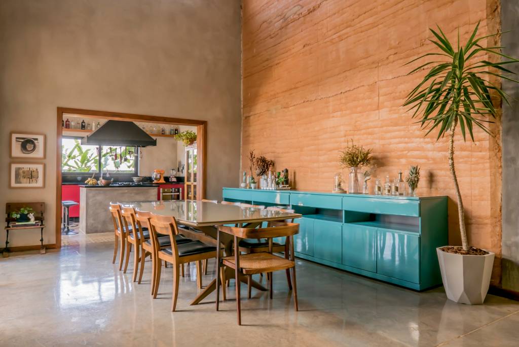 reforma-casa-veraneio-vira-endereço-principal-filipe-augusto-living-moveis-coloridos-taipa-de-pilao-sala-de-jantar-buffet imagem mostra buffet em laca azul com parede de taipa de pilão ao fundo e mesa de jantar