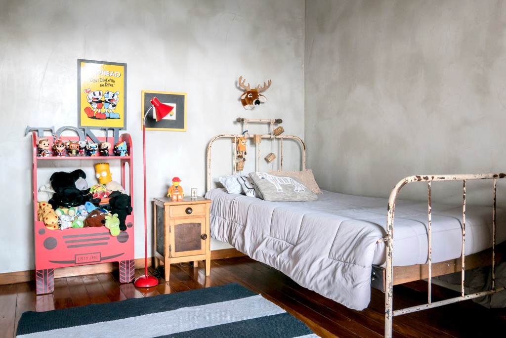 imagem mostra cama de ferro com cara de bem antiga, mesinha de apoio com gaveta e alguns brinquedos, quadros, luminária de chão na cor vermelha e tapete listrado sobre piso de madeira.