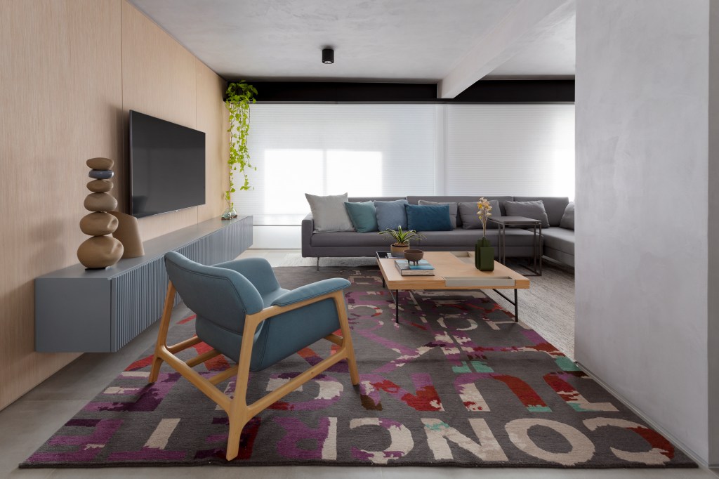 Sala de estar integrada com tapete colorido, sofá cinza e poltrona azul e painel de madeira