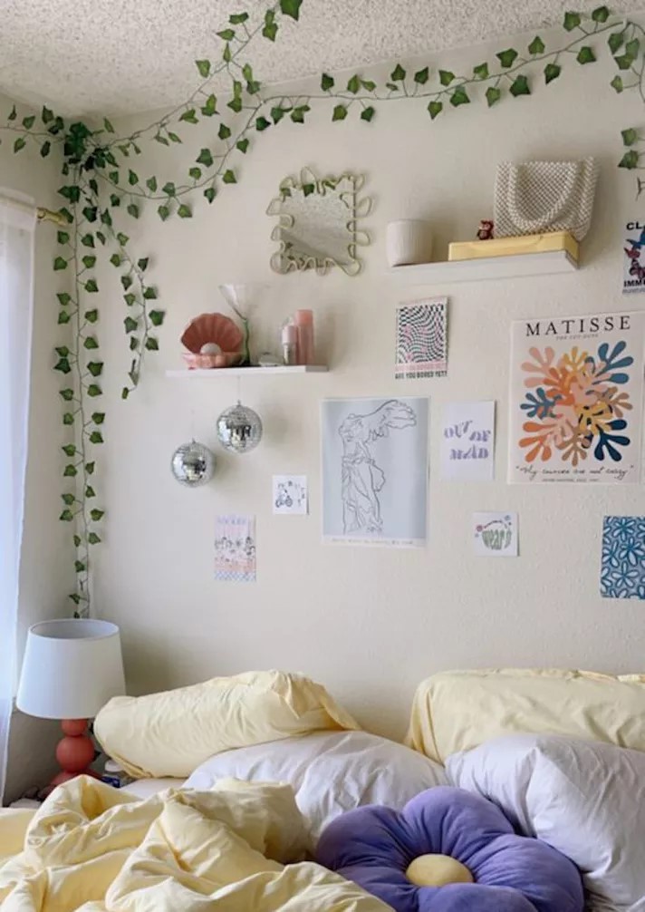 Quarto com cama coberta com almofadas em forma de flor e parede de cabeceira decorada por prints de obras de arte, prateleiras com objetos pessoais e uma trepadeira artificial no canto da parede.