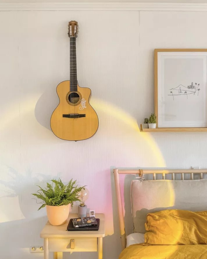 Quarto com mesa de cabeceira branca, iluminada por uma luz iridescente circular. Um violão pendurado na parede arremata o visual.