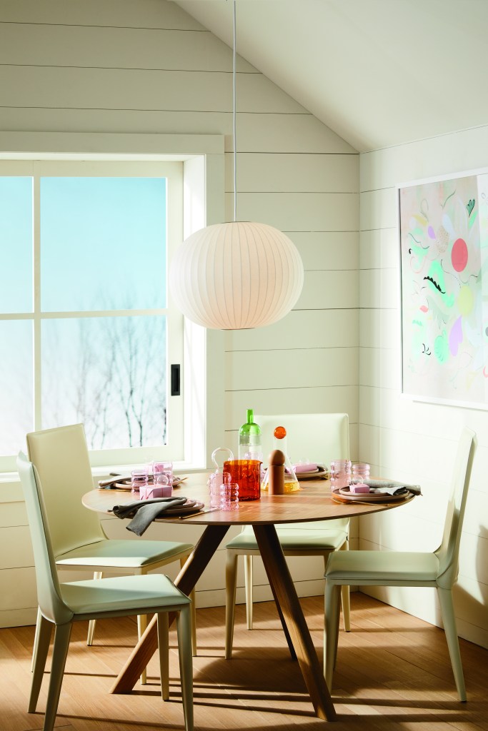 Mesa de jantar de quatro lugares, sob luminária esférica branca, em uma sala bem iluminada por uma janela.