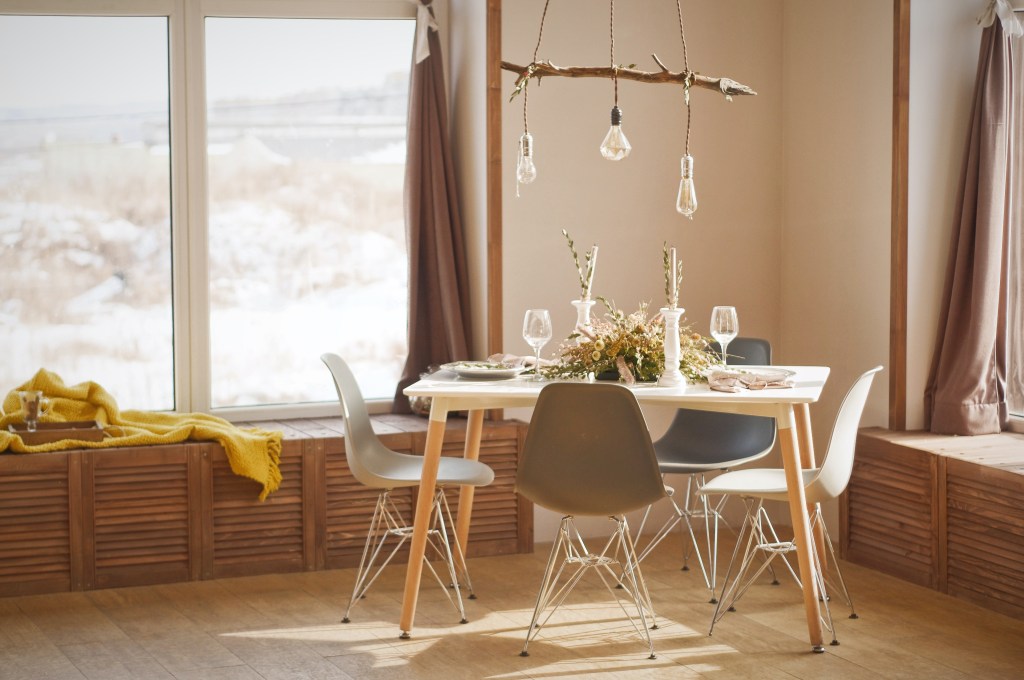 Mesa de jantar de quatro lugares, sob luminária de estrutura de madeira rústica em frente a uma janela.