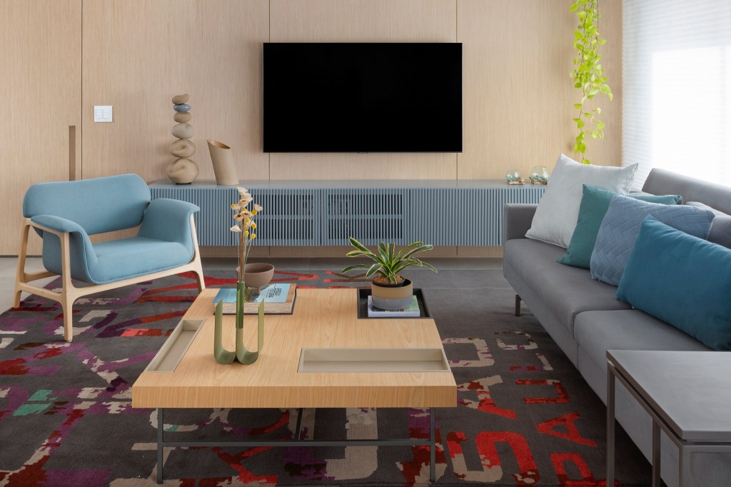 Living com painel de madeira, TV, tapete colorido e sofá cinza