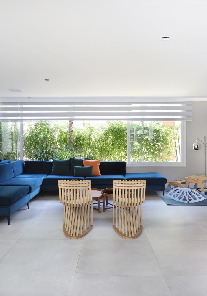Sala com sofá azul em L, janelas grandes voltadas à natureza e cadeiras em tons neutros