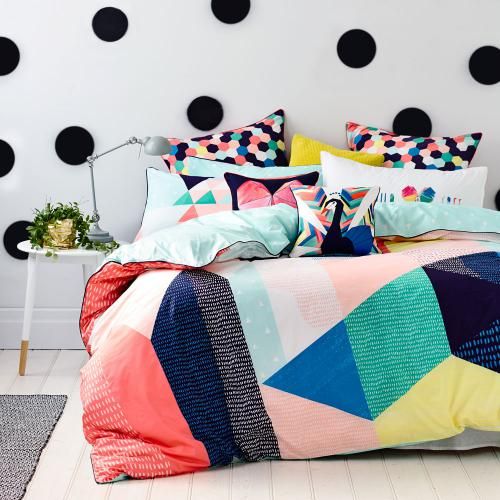 Quarto com cama decorada por edredom e fronhas coloridas em diversas formas geométricas. A parede da cabeceira é branca com bolas pretas uniformemente distribuídas.